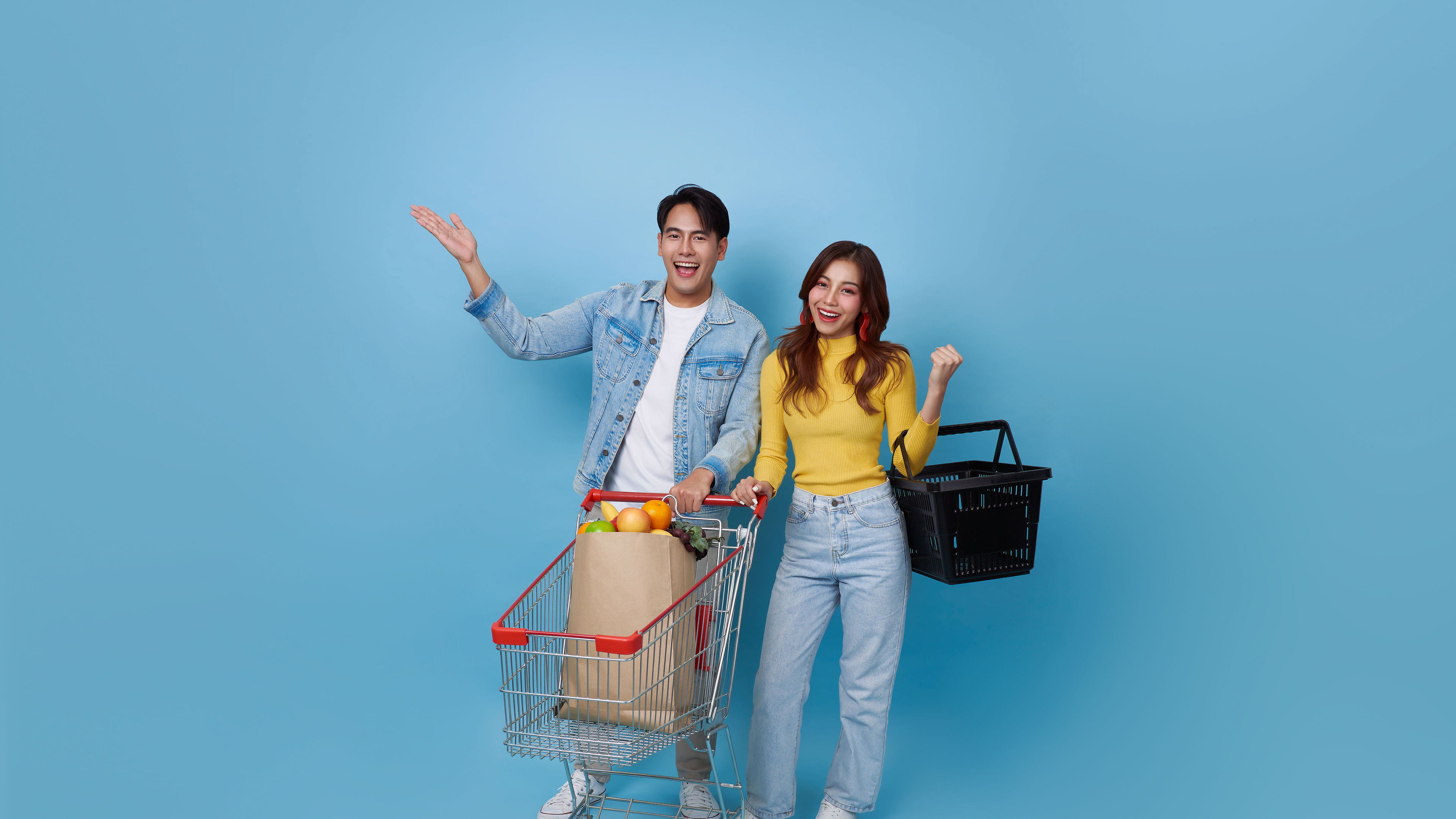 Young cheerful Asian couple with shopping trolley cart and holding shopping bag in a blue background
