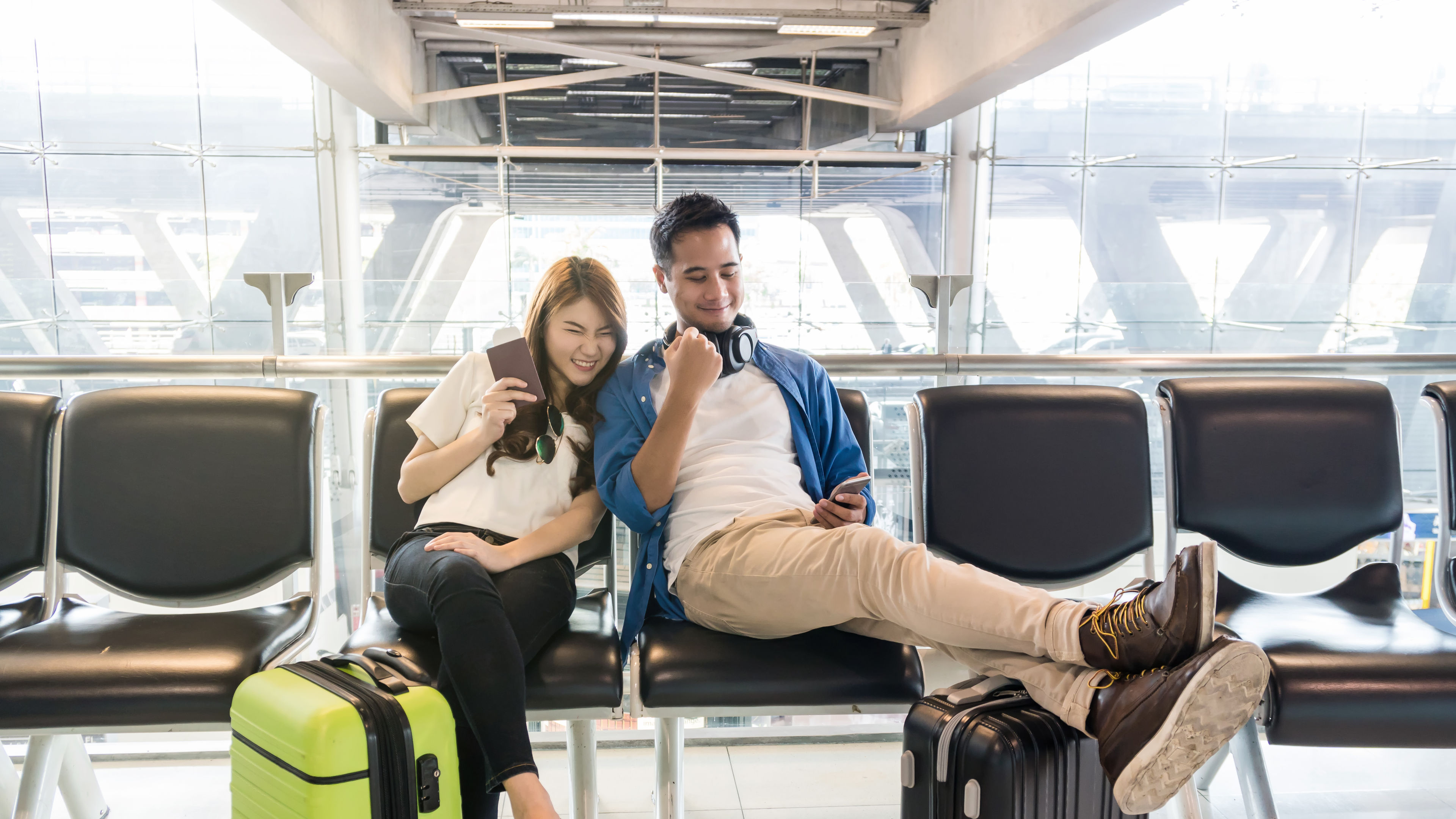 A young couple sitting in an airport terminal, smiling and relaxed, surrounded by luggage, with a modern glass background.