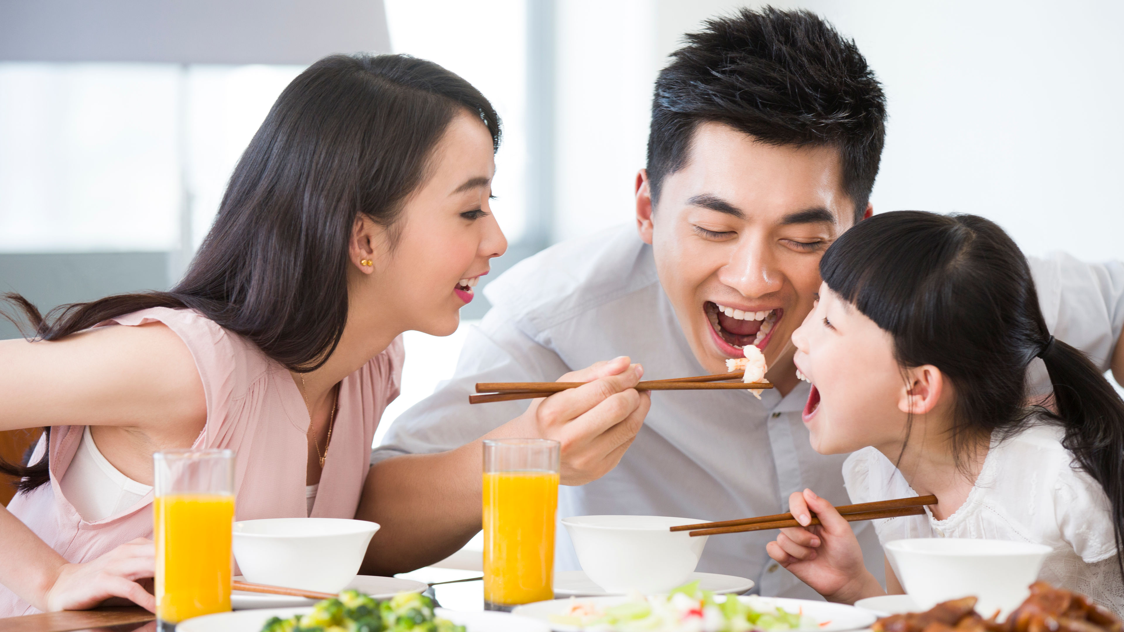 Masayang kumakain ang pamilyang Asyano, nakasuot ng mga damit na may kulay na liwanag, may katas sa mesa.