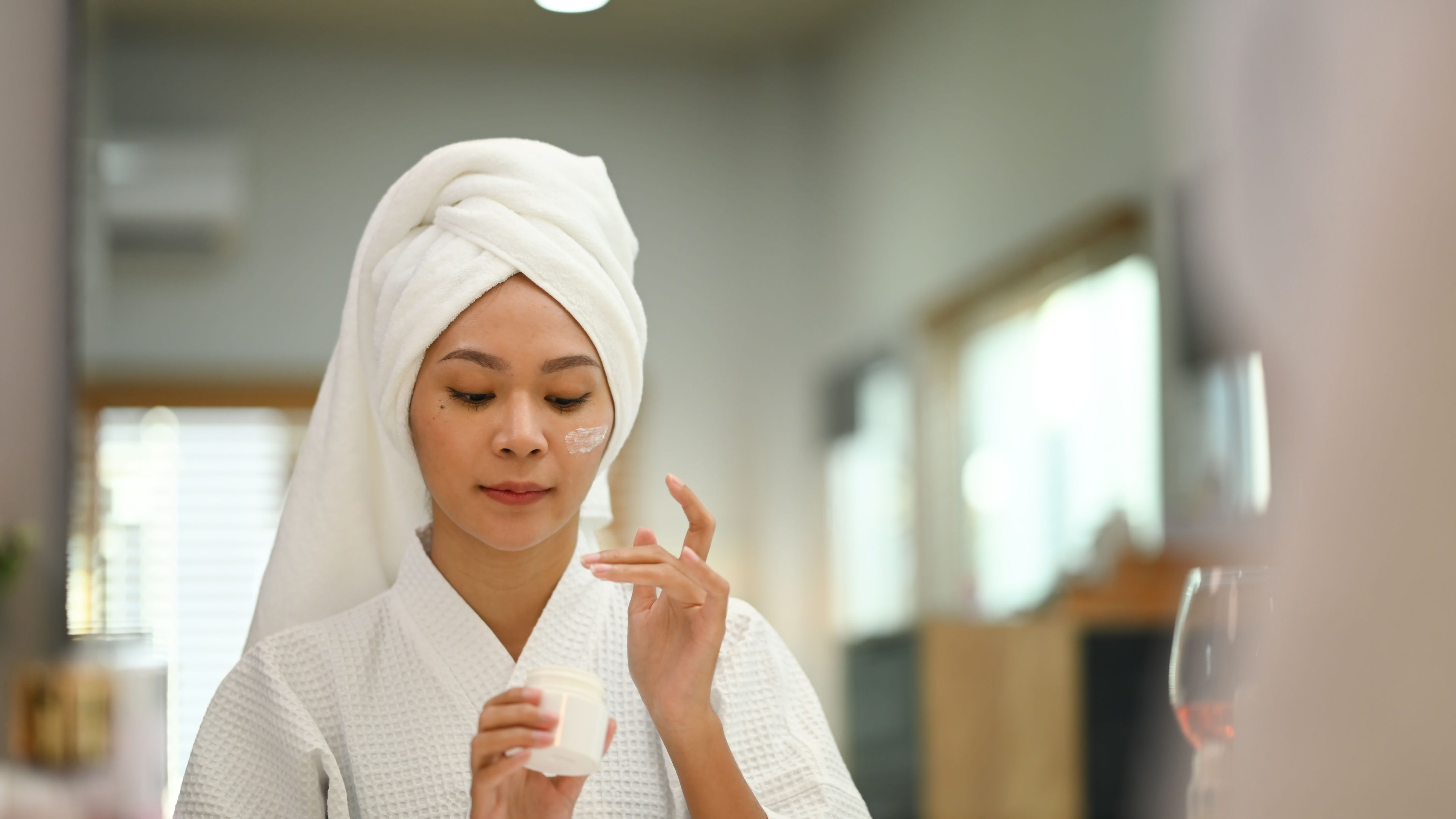 A beautiful woman in a bathrobe applying facial skincare cream. 