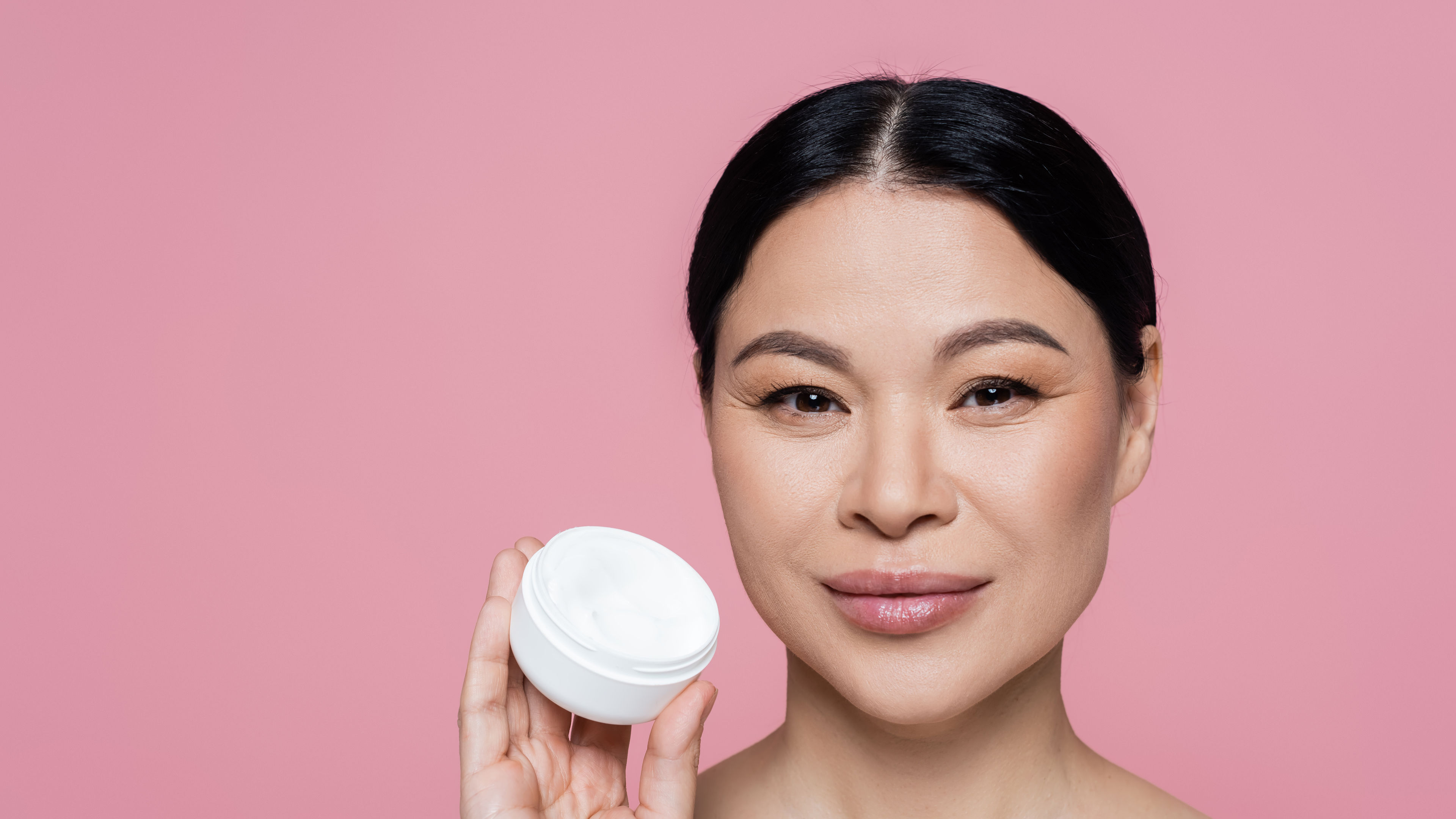 An Asian woman with bare shoulders is holding a cream, isolated on a pink background. 