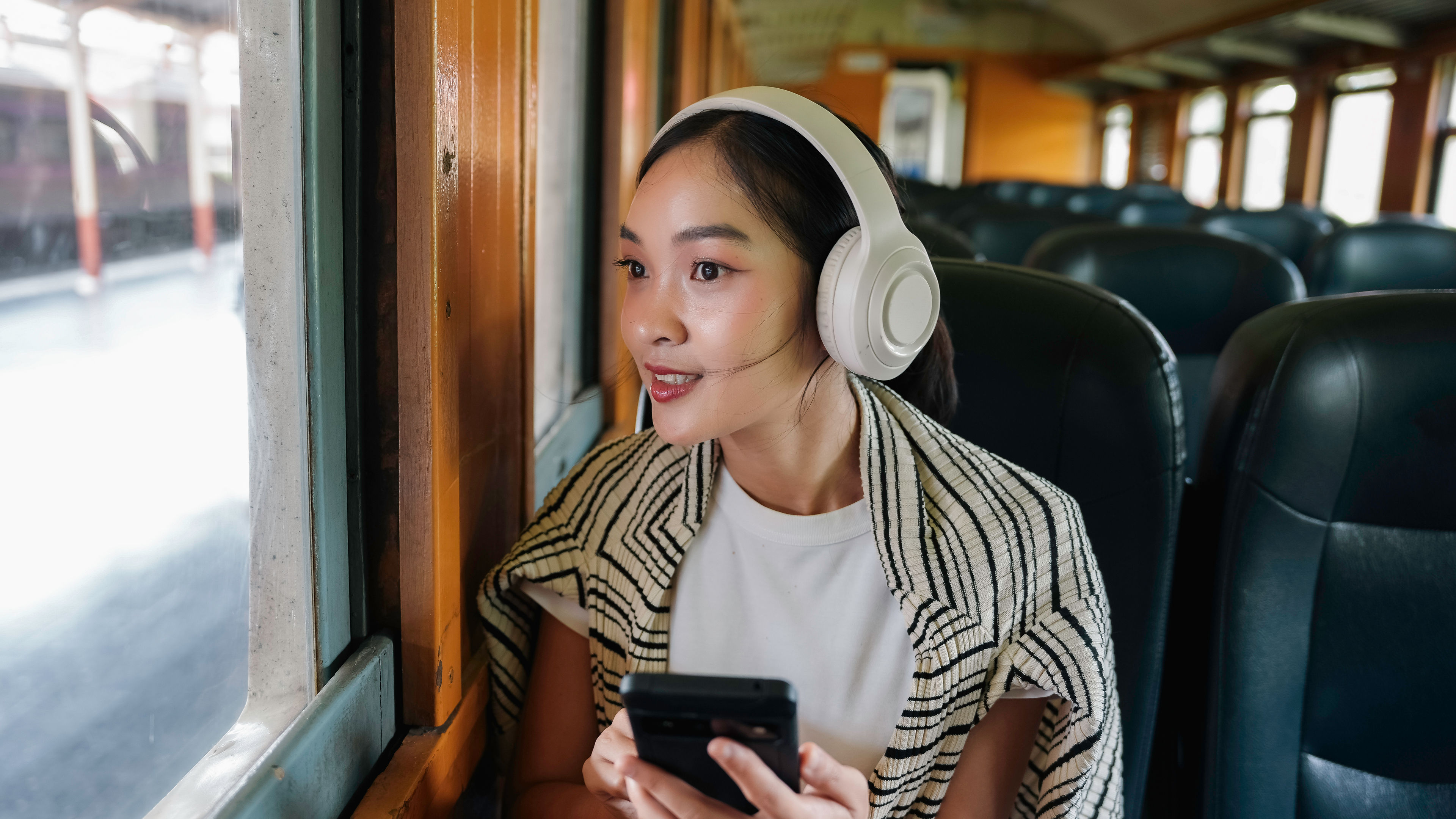 Young woman is commuting by train, listening to music on her headphones and holding her smartphone 